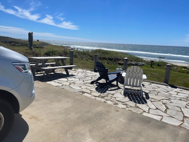 Seaside patio featuring an empty picnic table and two adirondack chairs overlooking the ocean, with waves gently breaking on the shore.