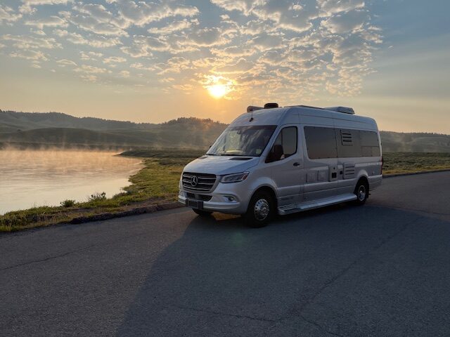 A sleek silver van parked by a lake at sunrise, with mist rising from the water and rolling hills in the background. Ideal for travel inspiration.