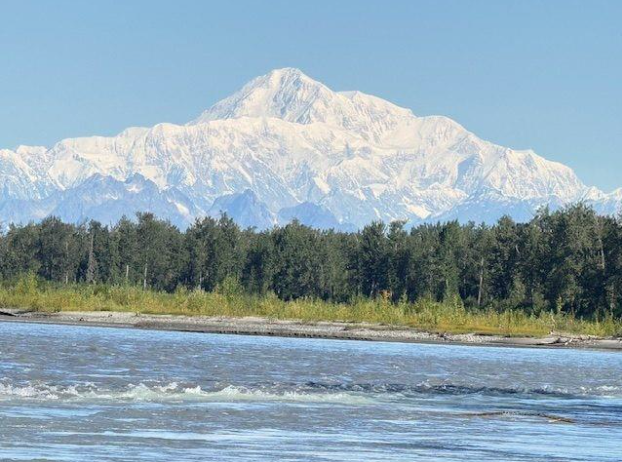 Snow-capped Denali rises majestically in the background, framed by a lush forest and a flowing river, showcasing Alaska's stunning natural beauty.