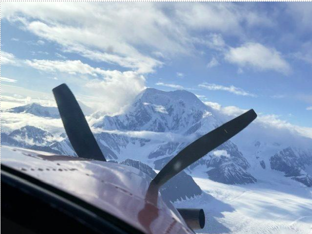 Aerial view from a plane cockpit, featuring a snow-covered mountain peak and blue sky with clouds. Highlights scenic flight over rugged terrain.