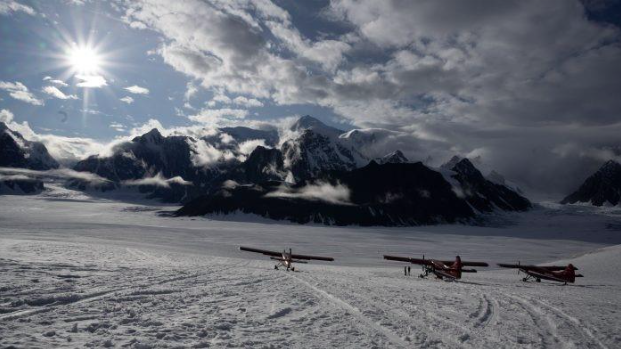 Two small red planes parked on a snow-covered glacier with rugged mountains and dramatic clouds in the background under a bright sun.