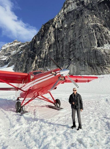A red bush plane parked on snow, with a rugged mountain backdrop. A person stands beside the aircraft, showcasing a remote winter landscape.