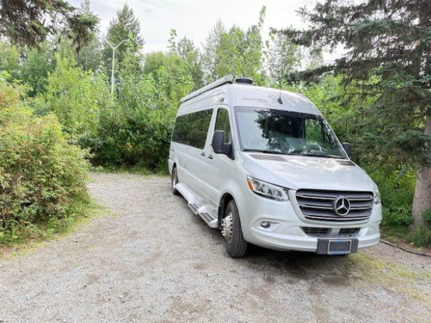 A silver Mercedes van parked on a gravel driveway, surrounded by lush green trees and foliage, suggesting a tranquil camping or travel setting.