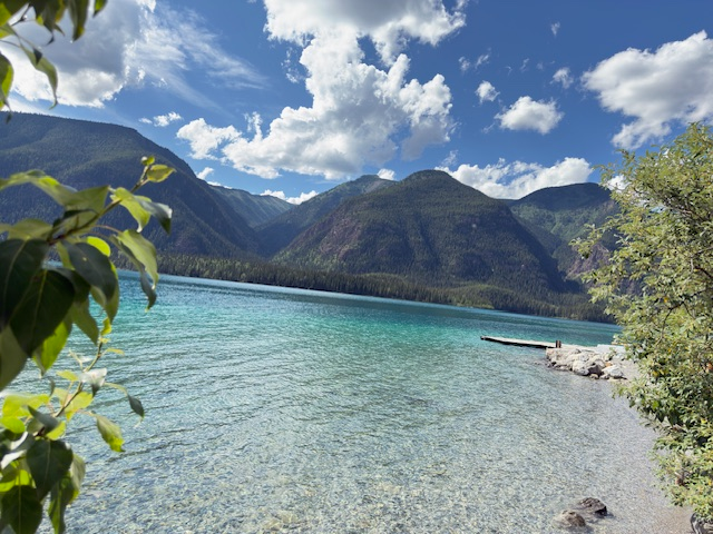 Clear turquoise water reflects mountains under a blue sky with fluffy clouds. A wooden dock extends into the lake, framed by lush greenery.