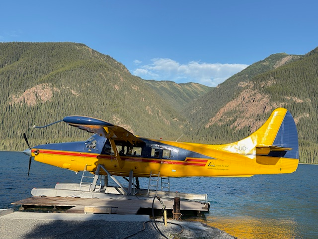 A yellow seaplane is docked by a serene mountain lake, surrounded by lush, forested hills under a clear blue sky.