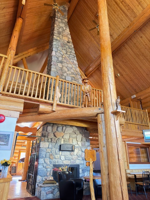 Spacious lodge interior featuring a stone fireplace and wooden beams, accented by a wooden statue on a balcony above. A cozy atmosphere.