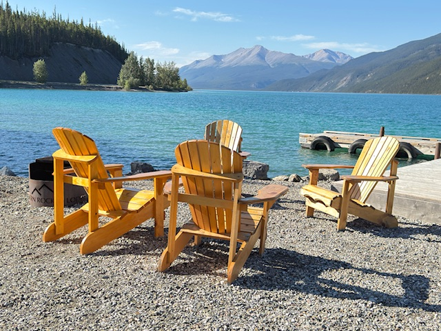 Adirondack chairs are lined up on a rocky shore by a clear blue lake, surrounded by mountains under a bright sky, inviting relaxation.