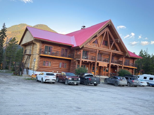 A large log cabin-style lodge with a red roof, surrounded by trees and parked vehicles. The setting suggests a rustic retreat in nature.