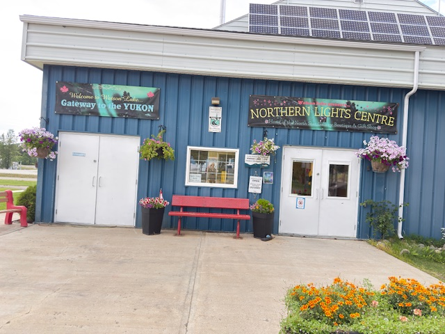 Welcome sign for the Yukon and the Northern Lights Centre, featuring bright flowers and a red bench in front of a blue building.