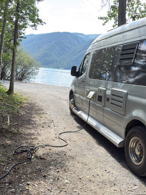 A camper van is parked near a lake, plugged into a power source, with mountains visible in the background. Ideal for outdoor adventures and eco-tourism.
